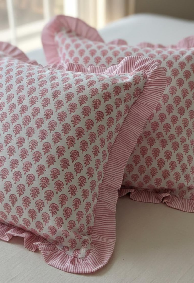 May include: Two decorative pillows with a white background and a repeating pink floral pattern. The pillows have a ruffled edge and a striped border in pink and white. The pillows are on a white surface.
