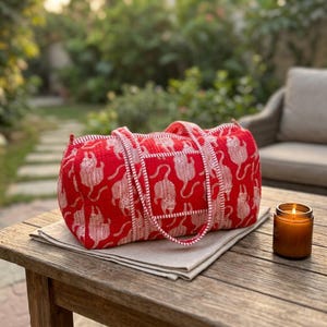 May include: A rectangular quilted duffel bag in bright red featuring a repeating white tiger pattern. The bag has two matching red and white striped fabric handles and a zippered top closure, showcasing a soft, textured fabric construction.