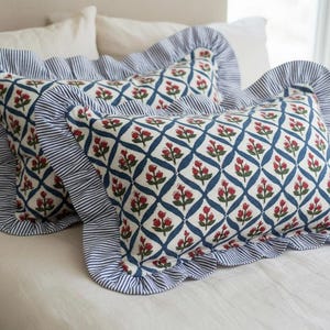 Op de afbeelding: Twee decoratieve sierkussens met een blauw-witte gestreepte ruchesrand. De stof heeft een witte achtergrond met een blauw ruitpatroon en rode bloemaccenten. De kussens liggen op een neutraal gekleurd bed.