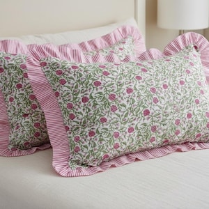 May include: Two decorative rectangular pillows with a floral pattern of pink flowers and green leaves on a white background. The pillows have a pink and white striped ruffle border. The pillows are on a white bed.