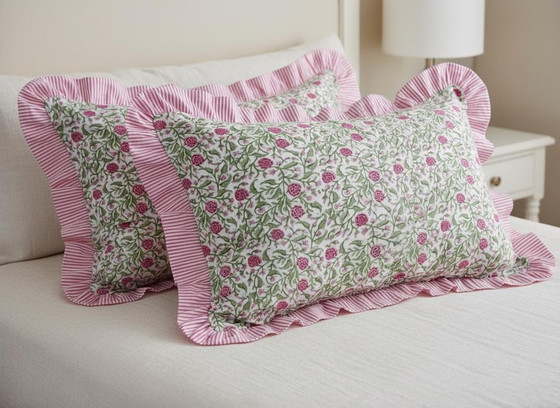 May include: Two decorative rectangular pillows with a floral pattern of pink flowers and green leaves on a white background. The pillows have a pink and white striped ruffle border. The pillows are on a white bed.