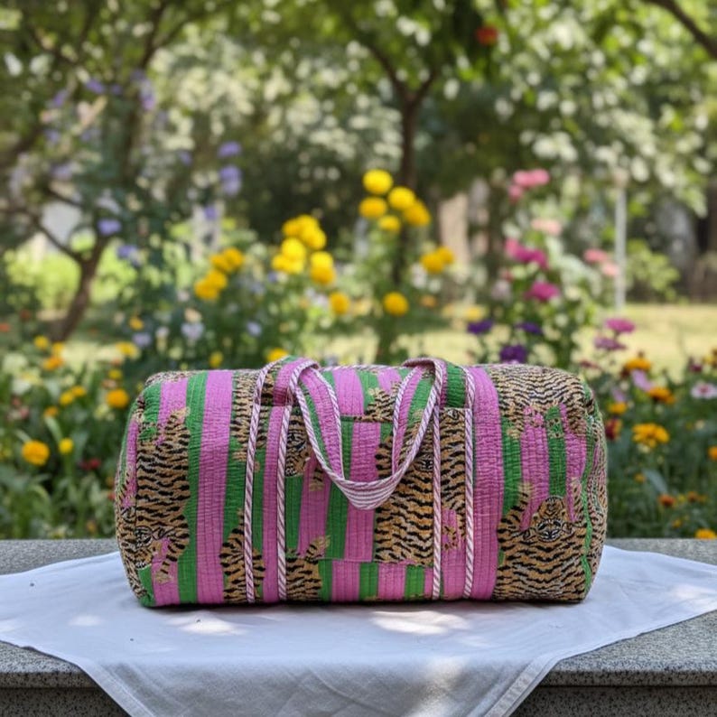 May include: A cylindrical duffel bag featuring a quilted texture with alternating vertical stripes of bright pink and green, decorated with a repeating tiger pattern throughout. The bag includes two matching fabric handles.