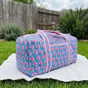 May include: A rectangular quilted duffel bag featuring a repeating floral pattern in shades of pink and green on a light blue background. The bag includes two fabric handles and a zipper closure, accented with pink and white striped trim.