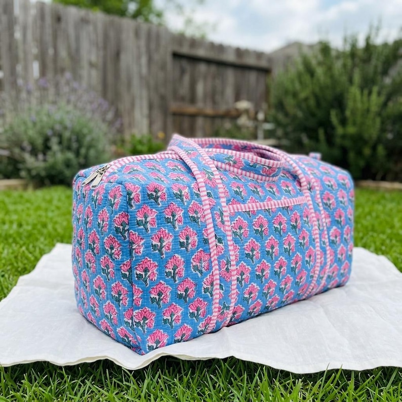 May include: A rectangular quilted duffel bag featuring a repeating floral pattern in shades of pink and green on a light blue background. The bag includes two fabric handles and a zipper closure, accented with pink and white striped trim.