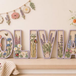 Könnte beinhalten: Ein Set aus Holzbuchstaben, die das Wort OLIVIA bilden, verziert mit geschichteten Blumen- und Naturmotiven. Die Buchstaben zeigen Mohnblumen, Lavendel, Gänseblümchen, eine Biene, einen Marienkäfer, einen Schmetterling, eine Gießkanne und einen Vogel.