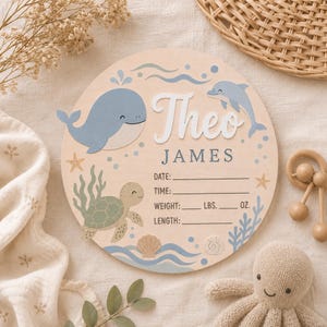 Puede incluir: Un letrero redondo de madera con un fondo de vetas de madera clara. Presenta una ballena, un delfín y una tortuga en azul y verde. El nombre "Theo James" está en blanco, con líneas para fecha, hora, peso (lbs, oz) y longitud.
