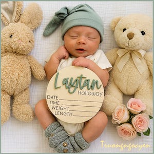 May include: A newborn baby wearing a green cap and gray knitted booties lies on a white surface, surrounded by a stuffed bunny and teddy bear. A wooden sign with the name "Laytan Holloway" and space for date, time, weight, and length is in front of the baby.