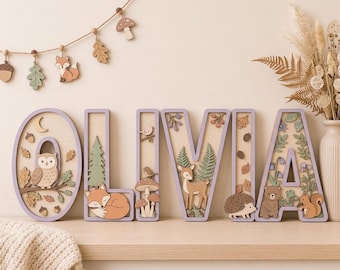 May include: A set of wooden letters spelling OLIVIA, each featuring layered forest-themed cutouts including an owl, fox, deer, hedgehog, bear, and squirrel, with light purple borders and natural wood tones, displayed as a decorative arrangement.