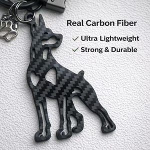 Puede incluir: Un llavero negro de fibra de carbono con forma de perro. El llavero está unido a un llavero plateado y a un dije en forma de pata. El texto de la imagen dice "Real Carbon Fiber", "Ultra Lightweight" y "Strong & Durable".