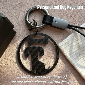 Puede incluir: Un llavero para perros de fibra de carbono negra con un diseño circular que presenta la silueta de un perro y un hueso. El llavero tiene un anillo plateado y una correa de cuero negro. El texto en la imagen dice: "Personalized Dog Keychain" y "A small everyday reminder of the one who's always waiting for you."