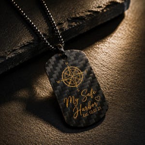Puede incluir: Un colgante de placa de identificación negro en una cadena de bolas a juego. El colgante presenta una textura de fibra de carbono con un gráfico de rosa de los vientos dorado y el texto grabado "My Safe Harbor" junto a un corazón y "Est. 12.01.21".