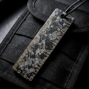 Puede incluir: Una regla rectangular de fibra de carbono con un patrón jaspeado en negro y gris con marcas de color dorado. La regla muestra incrementos numéricos de 0 a 15 centímetros a lo largo de su borde e incluye un orificio circular en la parte superior.