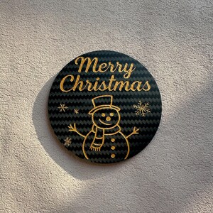 Puede incluir: Adorno navideño redondo y negro con letras doradas que dicen "Merry Christmas". El adorno presenta un diseño de muñeco de nieve dorado con sombrero, bufanda y copos de nieve.