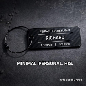 Puede incluir: Una etiqueta de equipaje negra de fibra de carbono con el texto "REMOVE BEFORE FLIGHT" y el nombre "RICHARD". La etiqueta también incluye "CF-90X28 | SERIES 01." y las palabras "MINIMAL. PERSONAL. HIS." y "REAL CARBON FIBER".