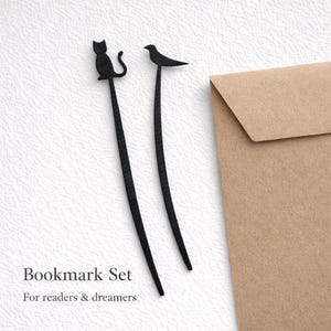 Puede incluir: Un juego de dos marcapáginas de metal negro. Uno presenta la silueta de un gato, el otro un pájaro. Los marcapáginas son largos y delgados, con las formas de animales en la parte superior. El texto "Bookmark Set" y "For readers & dreamers" está debajo.