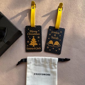 Puede incluir: Dos etiquetas de equipaje negras con correas amarillas. Una etiqueta dice "Merry Christmas" con un árbol y los nombres "Emma & Jack". La otra dice "Jessica & Sarah" con gorros de Papá Noel. Debajo hay una bolsa blanca con "ENJOYSMORE".