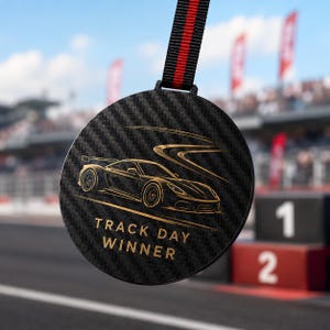 Peut inclure: Une médaille circulaire avec une texture en fibre de carbone noire présentant une illustration dorée d'une voiture de sport sur une piste sinueuse. Le texte "TRACK DAY WINNER" est imprimé en or sous la voiture. La médaille est suspendue à un ruban noir et rouge.