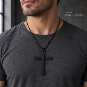 Puede incluir: Un colgante de cruz de gran tamaño con una textura de fibra de carbono oscura, suspendido de una cadena de metal negro. La cruz tiene un patrón tejido distintivo y se lleva sobre una camiseta gris oscuro. El texto LARGE STATEMENT SIZE es visible.
