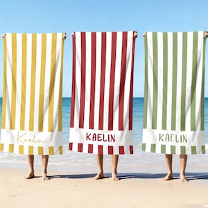 May include: Three beach towels with vertical stripes in yellow, red, and green, each with a white bottom border. The name "Kaelin" is printed on each towel. The towels are held up on a sandy beach with the sea in the background.