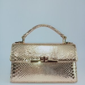 May include: A metallic gold handbag featuring a textured snakeskin pattern. The bag has a structured rectangular shape, a single top handle, and a polished gold-tone rectangular turn-lock closure centered on the front flap of the accessory.