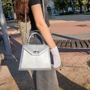 May include: Silver handbag with a snake-skin effect. The bag has a top handle and a shoulder strap. The clasp is silver. The bag is held by a person wearing a white glove. The background is a park setting with trees and a brick walkway.