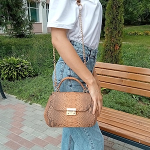 May include: A brown snakeskin handbag with a gold chain strap and a top handle. The bag has a rectangular shape with a gold clasp closure. The bag is being carried by a person wearing a white t-shirt and blue jeans.
