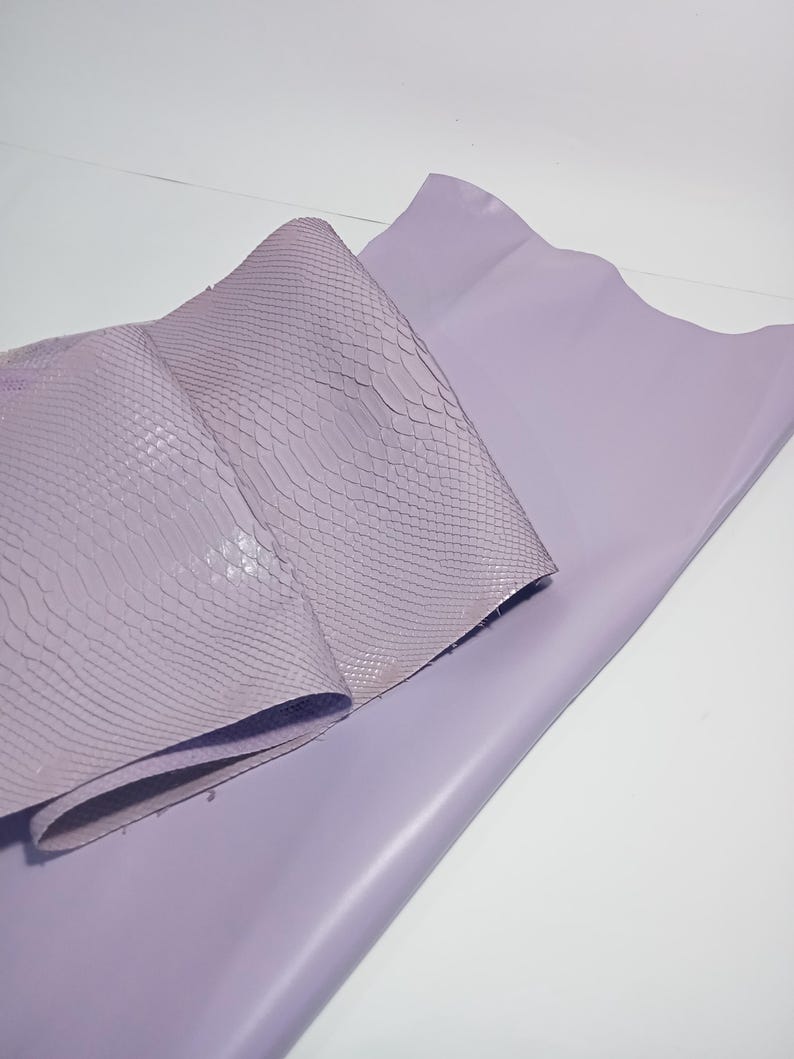 K&ouml;nnte beinhalten: Zwei Bl&auml;tter lavendelfarbenes Leder. Ein Blatt ist glatt und flach, w&auml;hrend das andere gefaltet und mit einem schuppenartigen Muster strukturiert ist. Das Leder wird wahrscheinlich f&uuml;r Bastelarbeiten oder Mode verwendet.