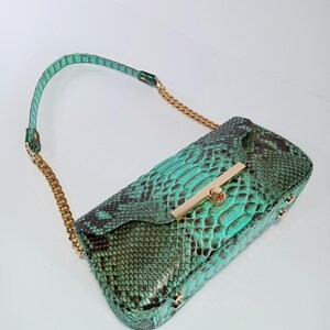 May include: A turquoise and black snakeskin handbag with a gold clasp and chain strap. The rectangular bag features a flap closure and small gold studs. The bag's design is structured.