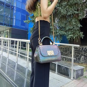 May include: A small, iridescent handbag with a snakeskin pattern. The bag has a gold chain strap and a top handle. The clasp is gold. The bag is held by a person wearing a green and black top and black pants.