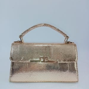 Op de afbeelding: Een metallic gouden handtas met een slangenleerachtige textuur. De rechthoekige tas is voorzien van een enkel handvat aan de bovenkant, een gestructureerde klepsluiting en een gepolijste goudkleurige metalen sluiting aan de voorzijde.
