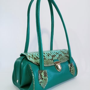 May include: A teal green handbag with a snakeskin flap and accents. The bag has two long, matching handles and a gold-colored clasp. The snakeskin pattern features shades of green and brown.
