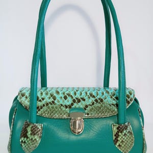 Puede incluir: Un bolso de mano turquesa con solapa y detalles de piel de serpiente. El bolso tiene un cierre plateado y asas turquesas a juego. El estampado de piel de serpiente presenta tonos verdes y azules. El bolso es de forma rectangular.