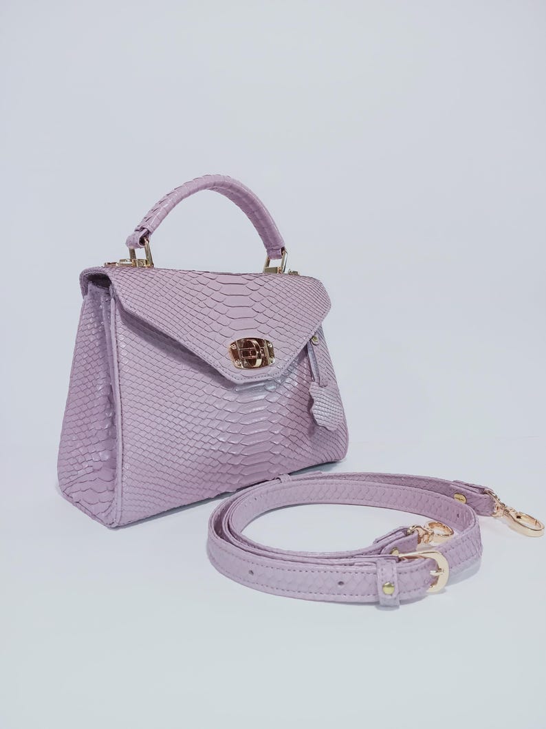 K&ouml;nnte beinhalten: Eine lavendelfarbene Handtasche mit einer strukturierten, schlangenlederartigen Oberfl&auml;che. Die Tasche hat einen oberen Griff, einen Klappverschluss und einen abnehmbaren Schulterriemen. Goldfarbene Hardware akzentuiert die Tasche.
