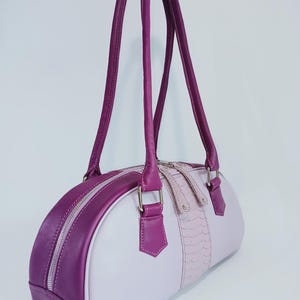 May include: A cylindrical handbag featuring a two-tone design with deep purple and light lavender panels. The bag includes long purple shoulder straps, silver-toned hardware, and a central vertical strip with a textured, scale-like pattern.