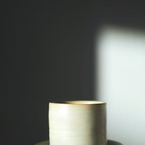 Può includere: Un set di tazza e piattino in ceramica fatto a mano. La tazza è cilindrica con un bordo leggermente svasato e il piattino è rotondo. Entrambi i pezzi sono di colore beige crema con un bordo marrone chiaro. Il set viene probabilmente utilizzato per bere caffè o tè.