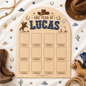 May include: An arched wooden milestone board featuring the text ONE YEAR OF LUCAS in blue, with a cowboy hat icon, a horse rider silhouette, and twelve empty square frames labeled from 1 month to 12 months for tracking baby growth.