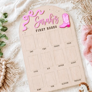 May include: Wooden milestone board with the text "Sarah's First Rodeo". The board has a pink bow and a pink cowboy boot. It features twelve rectangular spaces labeled with numbers, perfect for tracking a child's milestones.