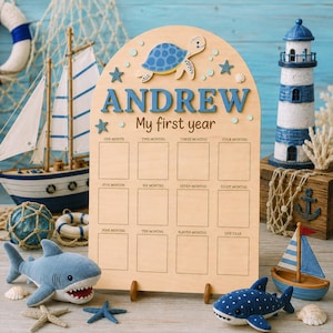 Puede incluir: Tablero de hitos de madera con el nombre "ANDREW" en letras azules, una tortuga azul y el texto "My first year". El tablero tiene espacios para fotos mensuales. Tema náutico con veleros de juguete, un faro y tiburones de peluche.