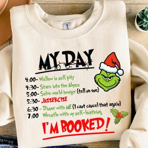 Peut inclure: Sweat-shirt crème avec le texte "MY DAY" et un dessin du Grinch portant un bonnet de Père Noël. Le sweat-shirt présente un emploi du temps d'activités, dont "Wallow in self pity" et "I'M BOOKED!"
