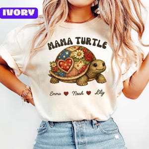 Puede incluir: Camiseta de color marfil con un diseño de tortuga colorida y el texto "MAMA TURTLE". El caparazón de la tortuga está decorado con patrones florales y de patchwork. Debajo de la tortuga están los nombres "Emma", "Noah" y "Lily".