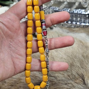 May include: A strand of yellow prayer beads with a silver tassel and decorative accents. The beads are a warm, amber colour and are rectangular in shape. The decorative accents include red and silver elements.