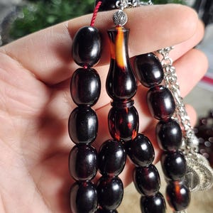 Op de afbeelding: Een donkerbruine gebedskralenset met ovale kralen. De kralen zijn geregen met een rood koord en hebben een decoratief middelpunt met zilveren accenten. De kralen worden in een hand gehouden.