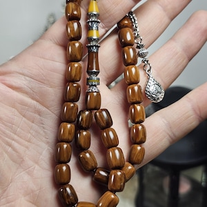 May include: A set of prayer beads with brown, rectangular beads. The beads are strung together with a silver chain and decorative accents. The focal point features a yellow and silver design. A silver teardrop charm hangs from the chain.