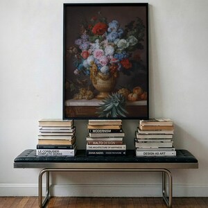 May include: A framed still life painting of a floral arrangement in a decorative vase with fruit, displayed above a black tufted bench with metal legs. Three stacks of books are arranged on the bench, featuring titles like Modernism, Aalto Design, and Design as Art.