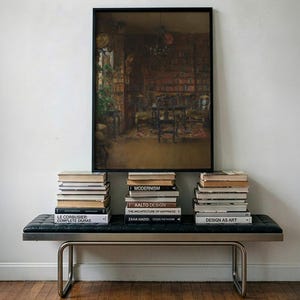 May include: A rectangular framed painting depicting a dark, detailed library interior with bookshelves and furniture, centered above a black tufted leather bench with metal legs, flanked by three stacks of various hardcover books.