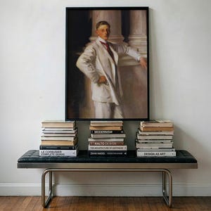 May include: A framed portrait painting of a person in a white suit with a red tie, displayed above a black tufted bench with a metal frame. Three stacks of books are arranged on the bench, featuring titles like Modernism and Zaha Hadid.