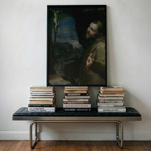 May include: A rectangular black-framed painting depicting a bearded figure in a brown robe looking toward a crucifix, mounted above a black tufted leather bench with a tubular metal frame, flanked by three stacks of various hardcover books.