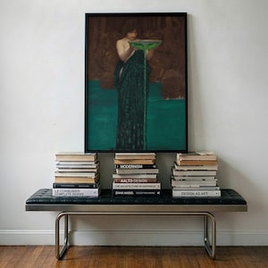 May include: A framed rectangular wall art print depicting a figure in a dark green robe holding a bowl with green liquid pouring out, positioned above a black leather bench with metal legs and three stacks of books including titles like Aalto Design.