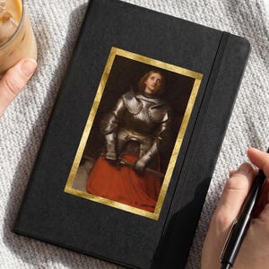 May include: A black notebook featuring a rectangular print of a person in silver plate armor and a red skirt, framed in gold. The notebook has a black elastic closure and is positioned next to a glass of iced coffee and a black ballpoint pen.