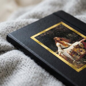 May include: A black hardcover journal featuring a rectangular inset print of a painting depicting a person with long red hair in a white dress, framed by a metallic gold border. The cover has a textured finish and a clean, rectangular shape.